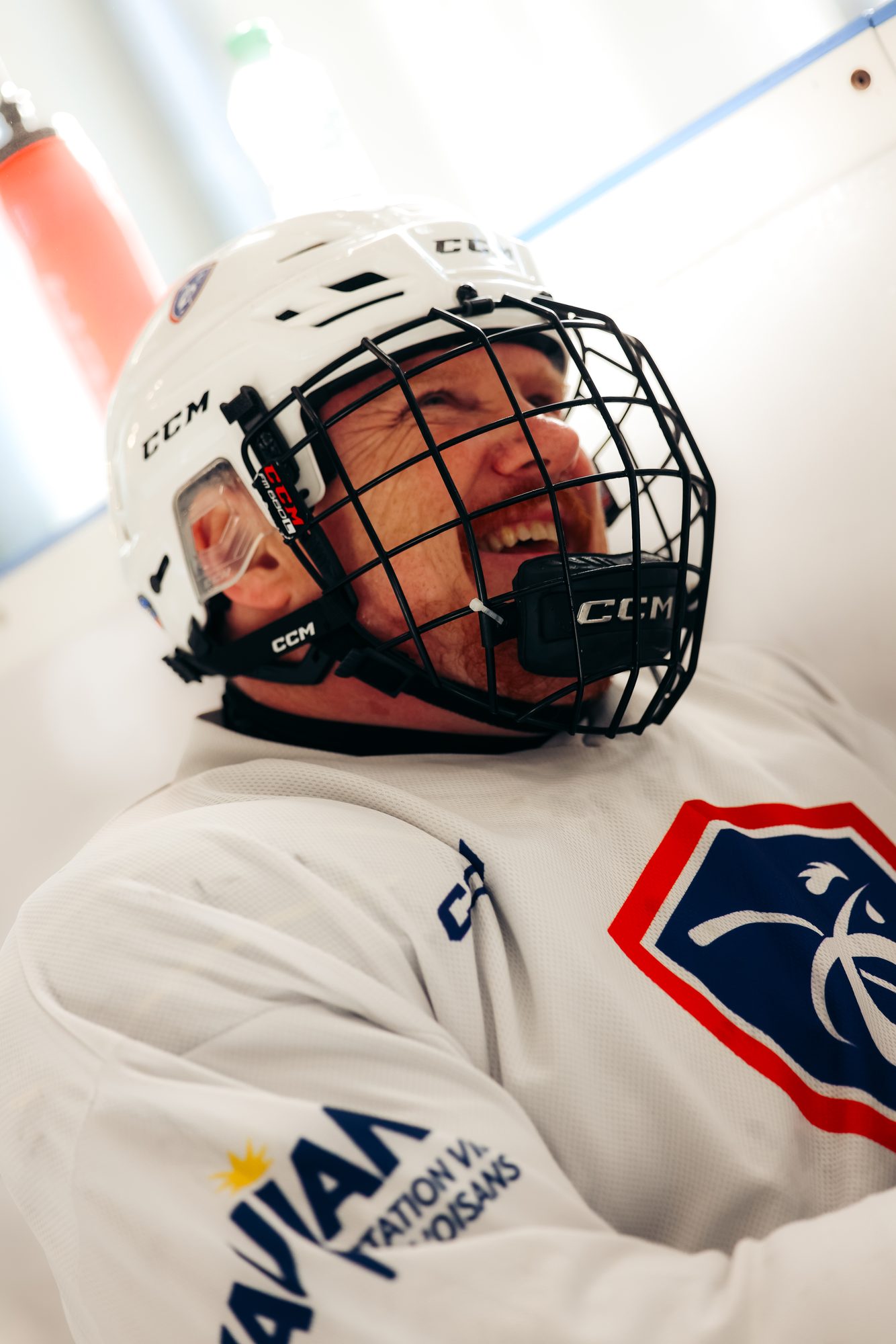 Gros plan sur un joueur de l'équipe de France en maillot blanc, casque blanc à grille noire, hilare, adossé à la bande sur sa luge