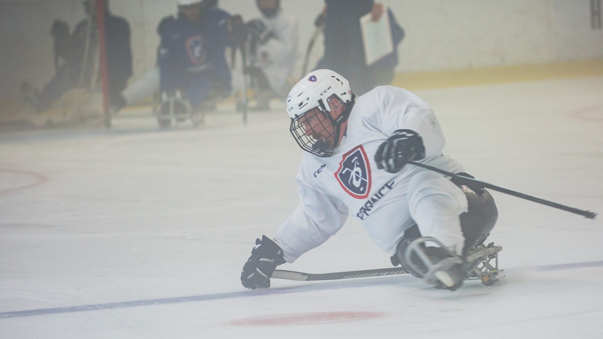 Un joueur de para hockey sur glace de l'équipe de France penché sur sa luge, une crosse dans chaque main