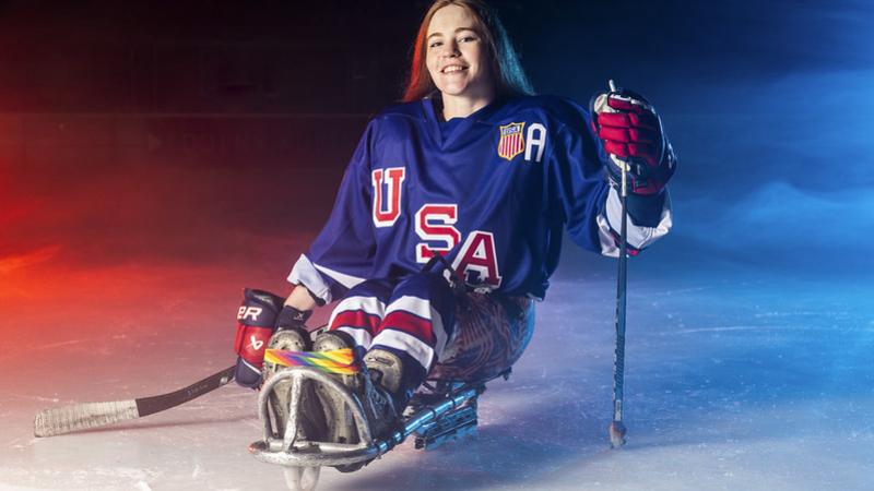Une joueuse de para hockey sur glace de l'équipe des États-Unis, assise sur sa luge, une crosse à la main : on distingue le baquet, les lames, les gants et les coudières