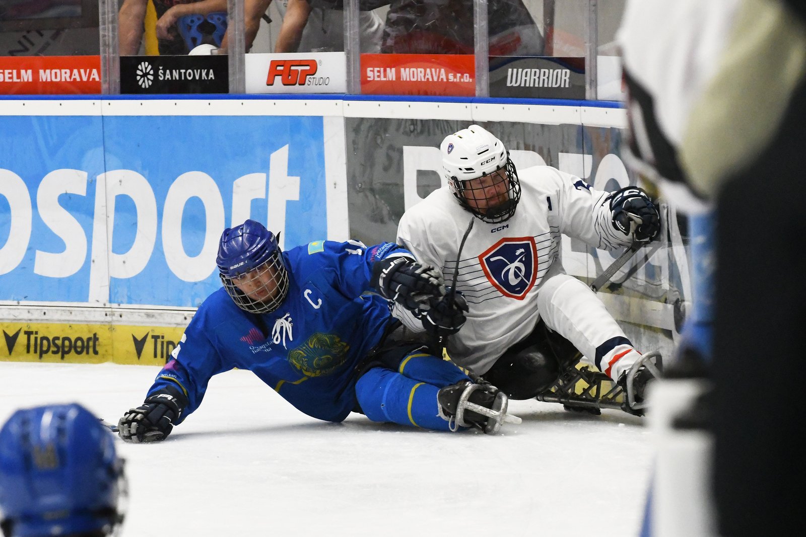 Le capitaine kazakh en maillot bleu, le C sur la poitrine, éjecté de sa luge et couché sur la glace, tenu contre la bande par un joueur français en blanc
