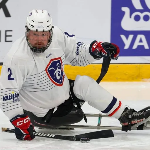 Yannick Jouenne en action sur sa luge de para hockey sur glace, en maillot blanc de l'équipe de France, le palet devant lui